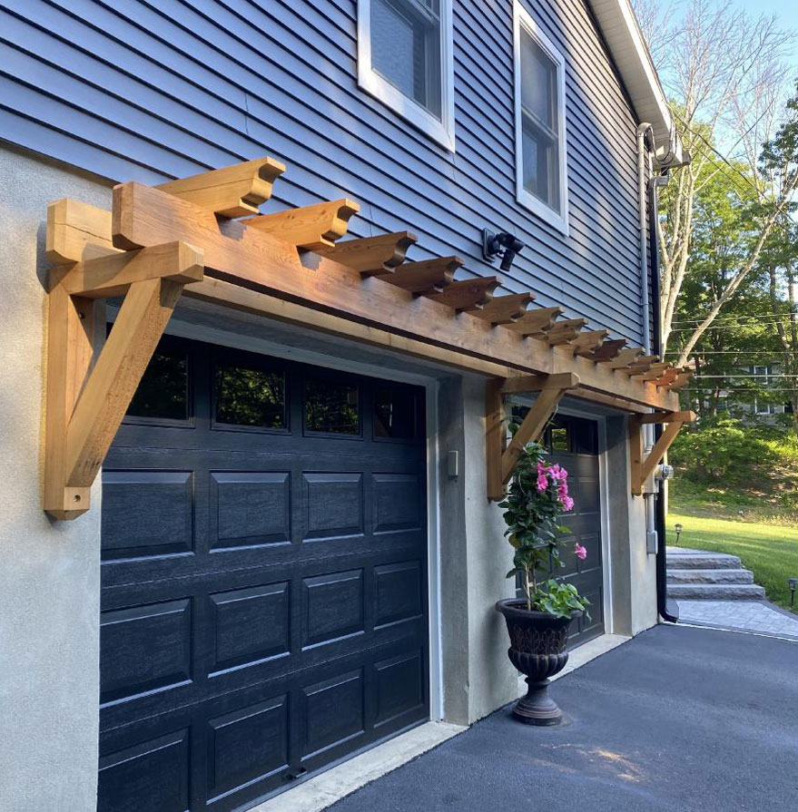 Beautiful wooden rafter tails support a pergola over a garage, enhancing the home's exterior with natural charm and architectural interest against a backdrop of lush greenery.
