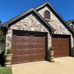 Charming brick garage with dual wooden doors, framed by elegant planters; featuring gable vents that enhance ventilation and architectural appeal against a clear blue sky.
