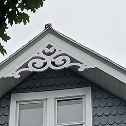 Elegant gable pediment featuring intricate scrollwork, enhancing the architectural charm of a home with gray scalloped siding and a cloudy sky backdrop. Perfect for detailed home design.