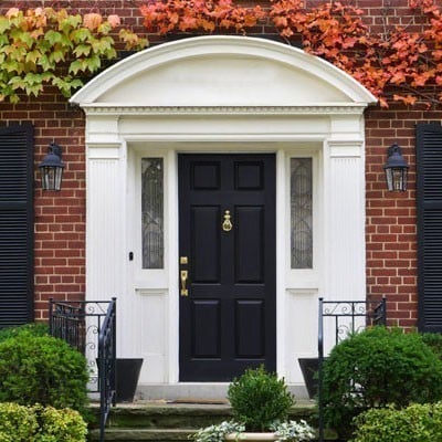 Elegant entryway featuring a black front door with detailed millwork, surrounded by tasteful landscaping and classic brick architecture, enhanced by vibrant autumn foliage.